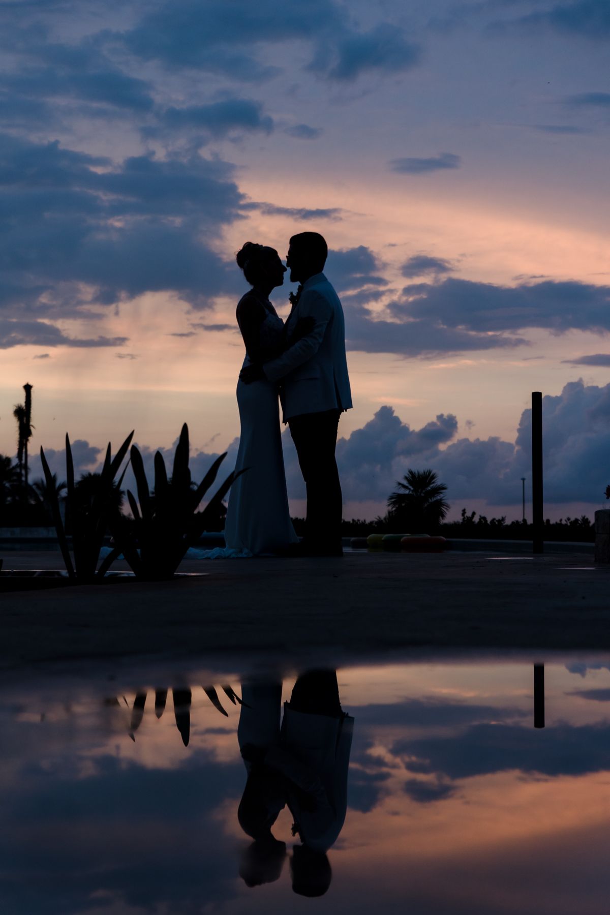 Silhouette of bride and groom kissing at sunset with dramatic purple sky reflected in water below.