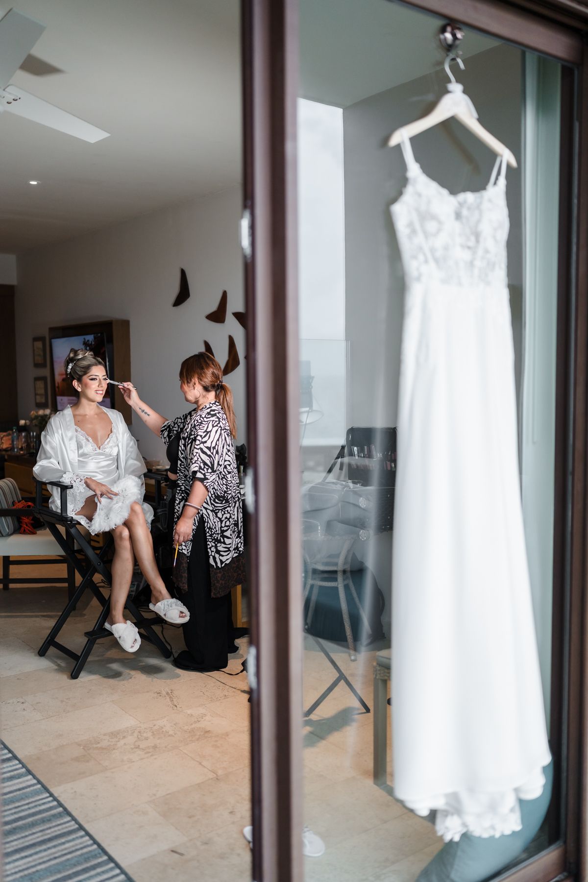 Bride getting makeup done in a white robe while her wedding dress hangs elegantly in the foreground.