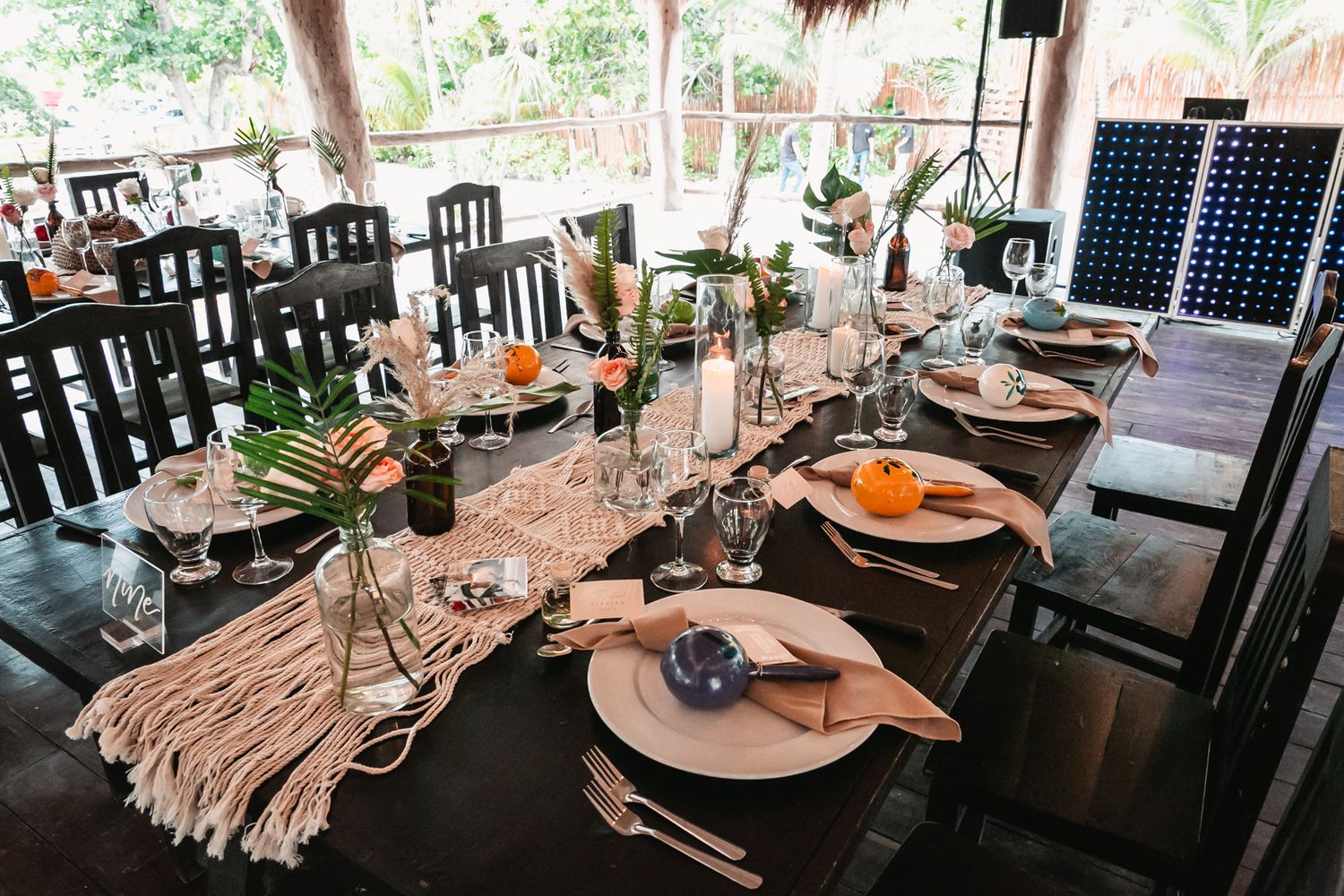 Rustic outdoor wedding reception table set with macrame runner, tropical flowers, and candles.