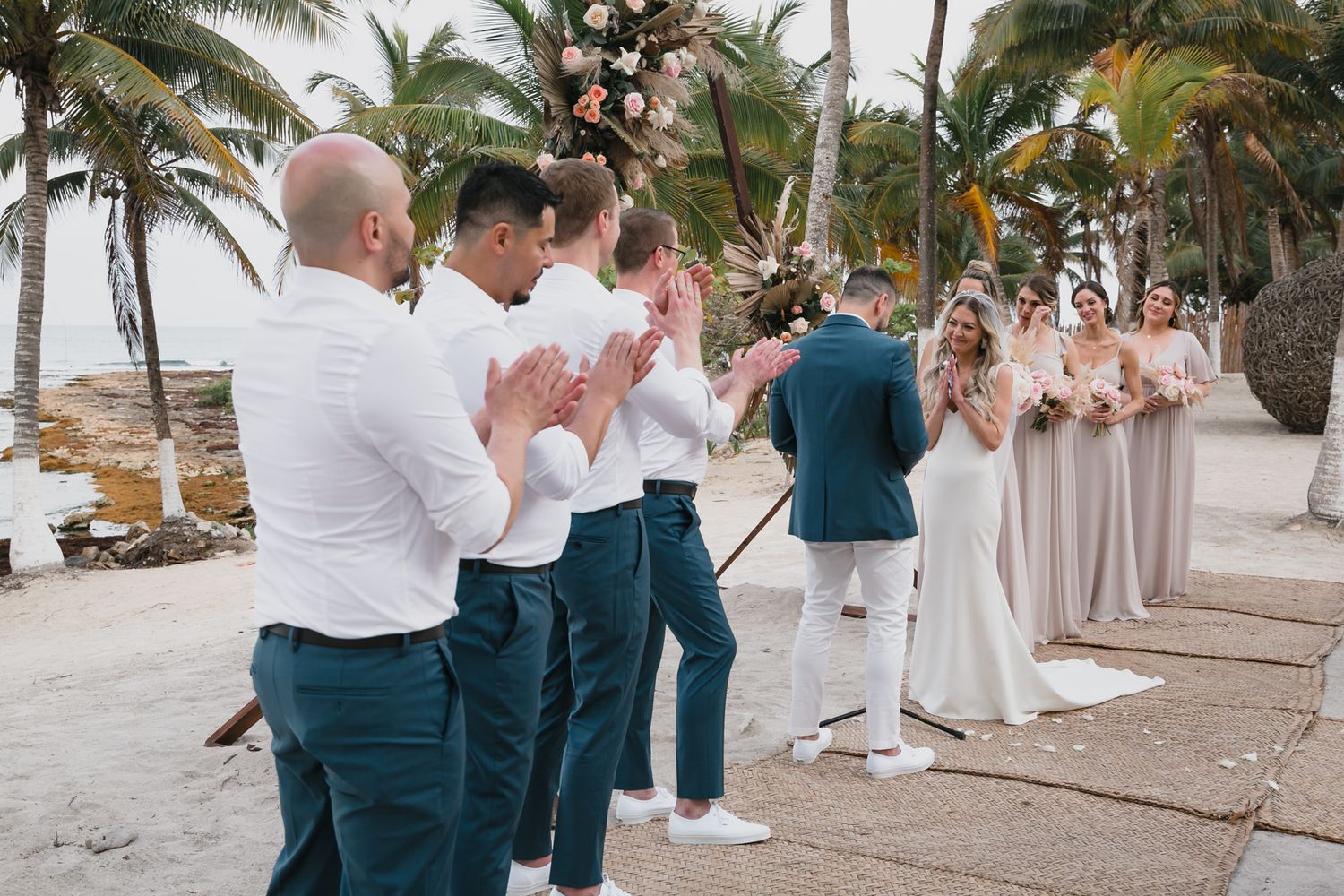 Beach wedding ceremony with groomsmen clapping as bride and groom exchange vows near palm trees.