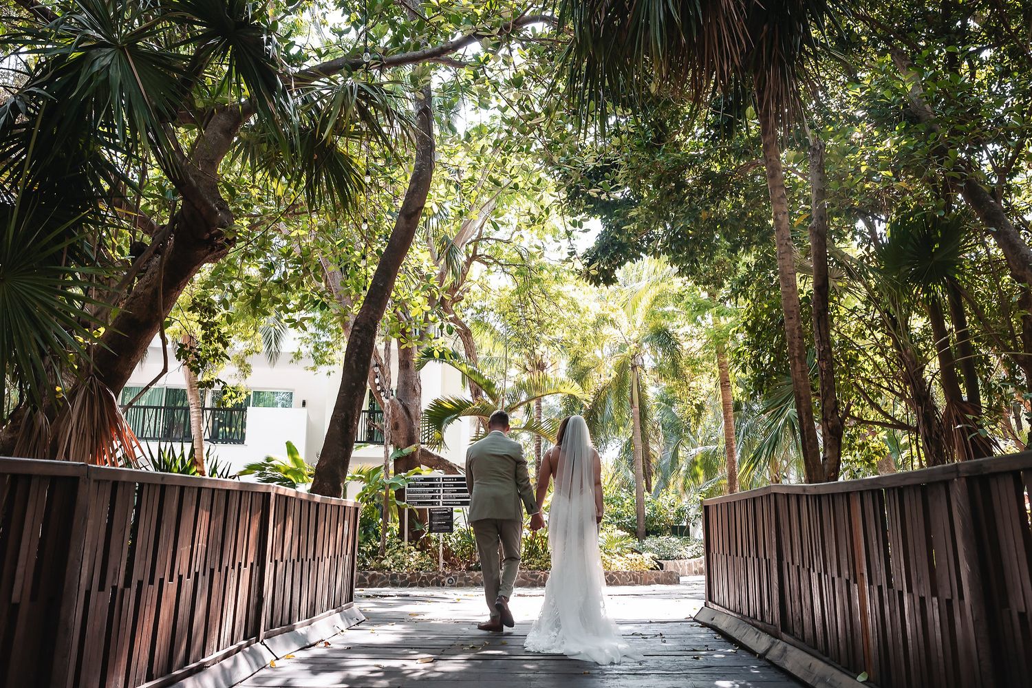 Bride and groom walking hand in hand across a wooden bridge surrounded by lush tropical palm trees at an outdoor wedding venue.