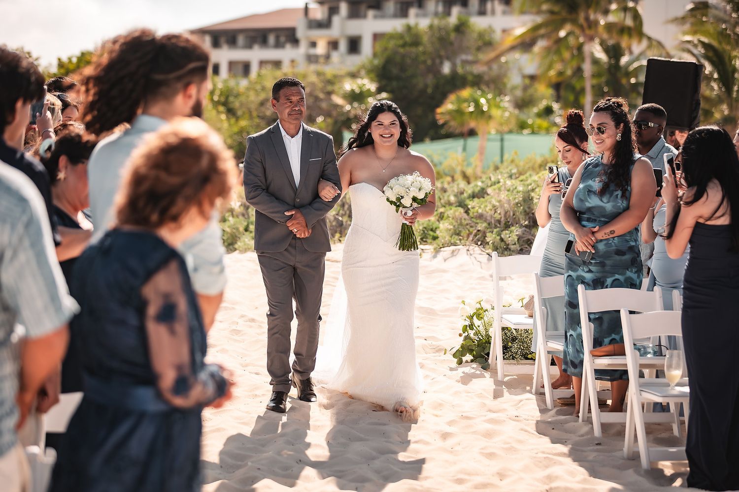 Bride walks down sandy beach aisle with escort at outdoor wedding ceremony, guests watching in tropical setting.