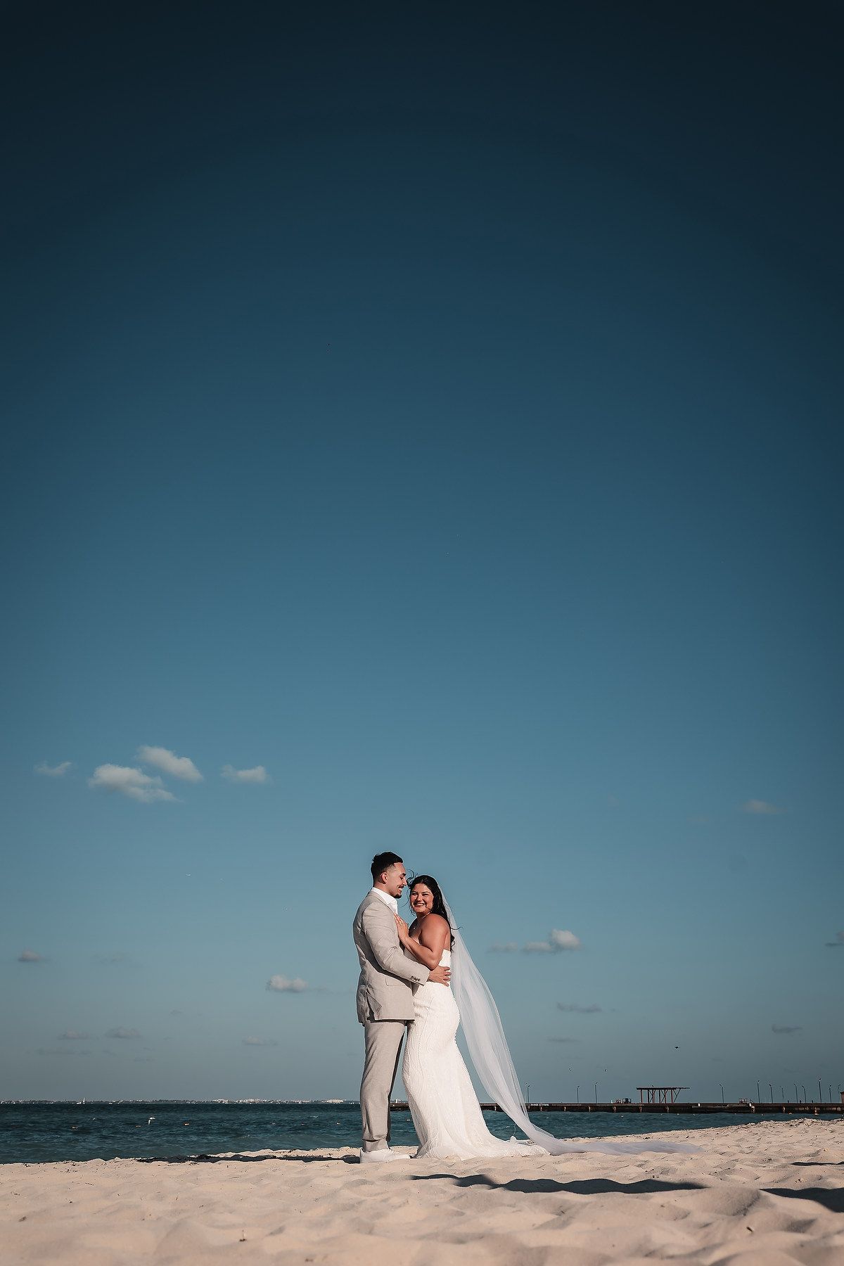 Bride and groom embracing on white sand beach under deep blue sky during destination wedding.