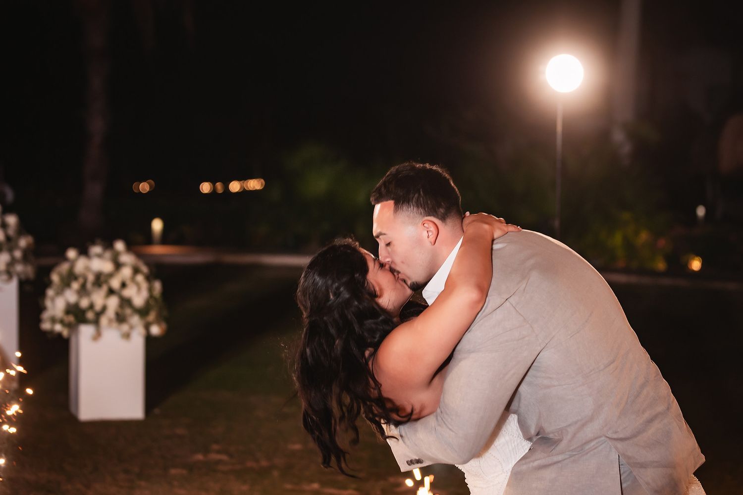 Bride and groom share a romantic kiss at night with fairy lights and white floral arrangements in the background.