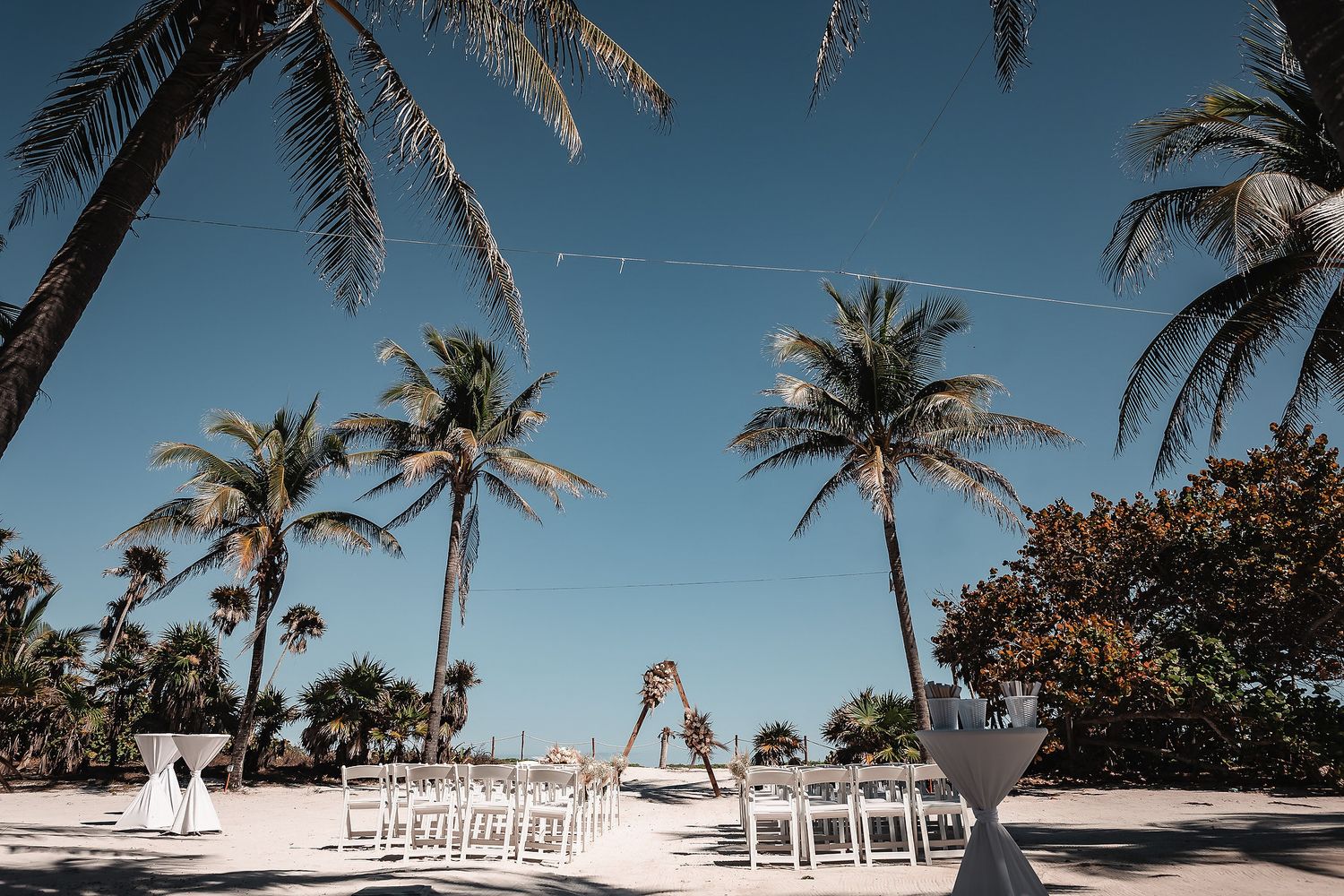 Beach wedding ceremony setup with white chairs and palm trees under a clear blue sky.