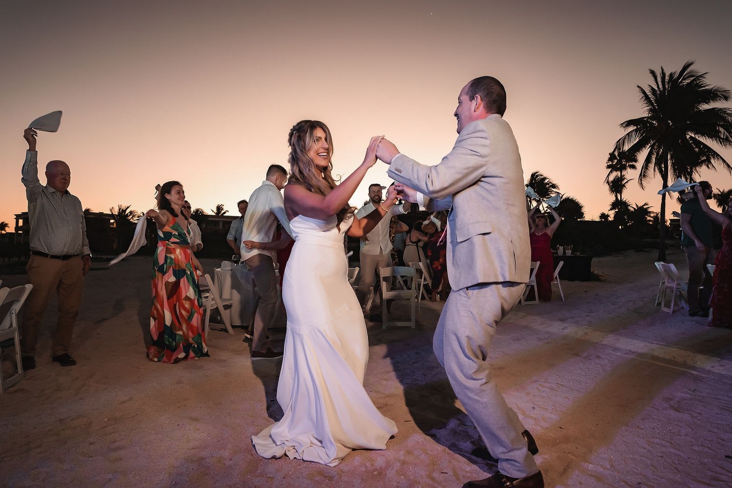 Bride and groom share first dance at outdoor beach wedding reception during golden sunset with guests celebrating in background.