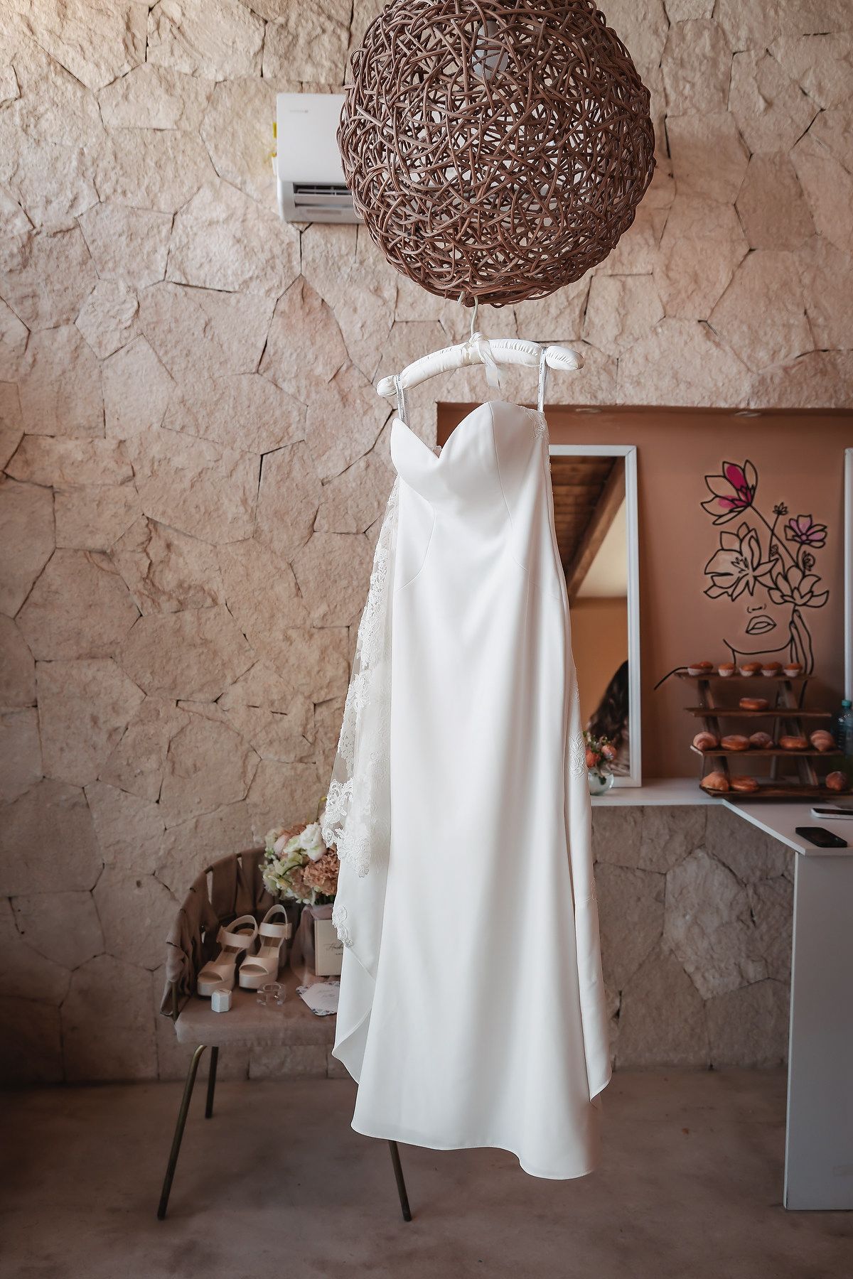 White wedding dress hanging on a decorative wicker pendant light in a rustic stone-walled bridal suite.