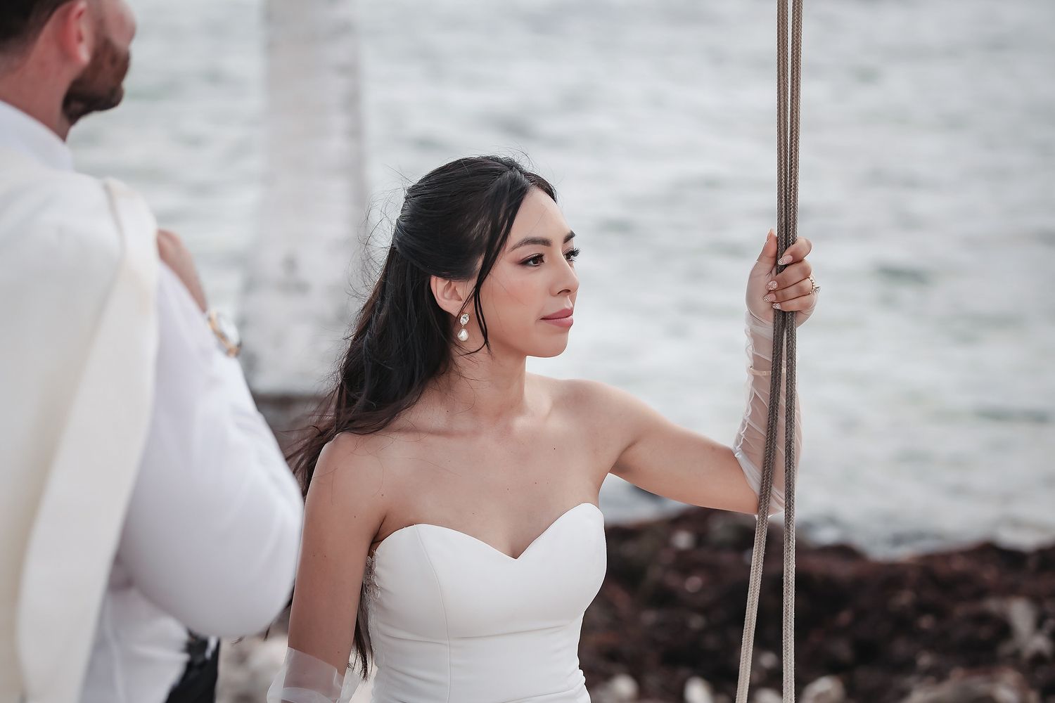 Bride in strapless white wedding gown holding a rope near the ocean shore on her wedding day.