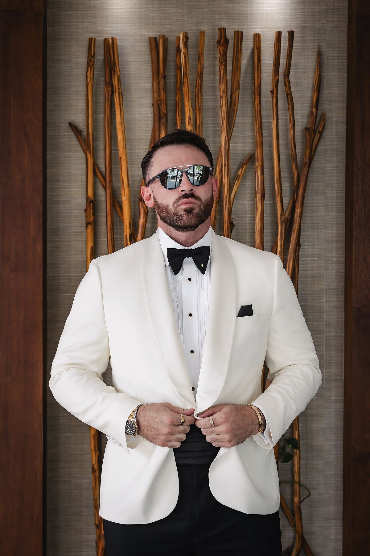 A confident groom in a white tuxedo jacket and bow tie adjusts his lapels, wearing mirrored sunglasses indoors.