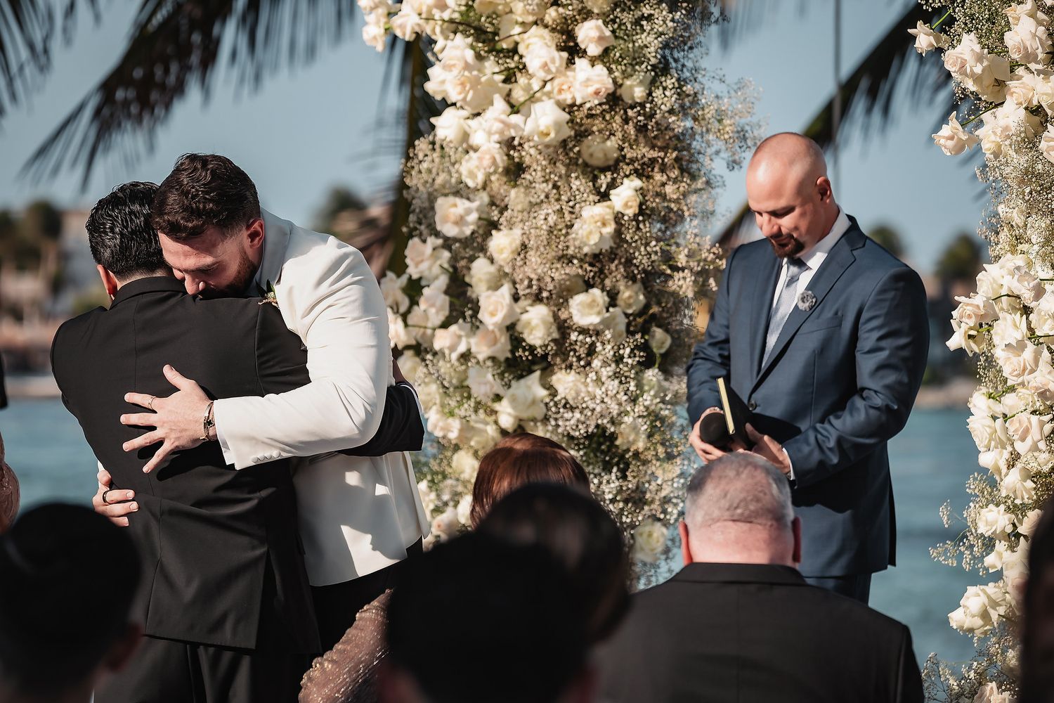 Two grooms share an emotional hug at outdoor wedding ceremony near floral arch with white roses and palm trees.