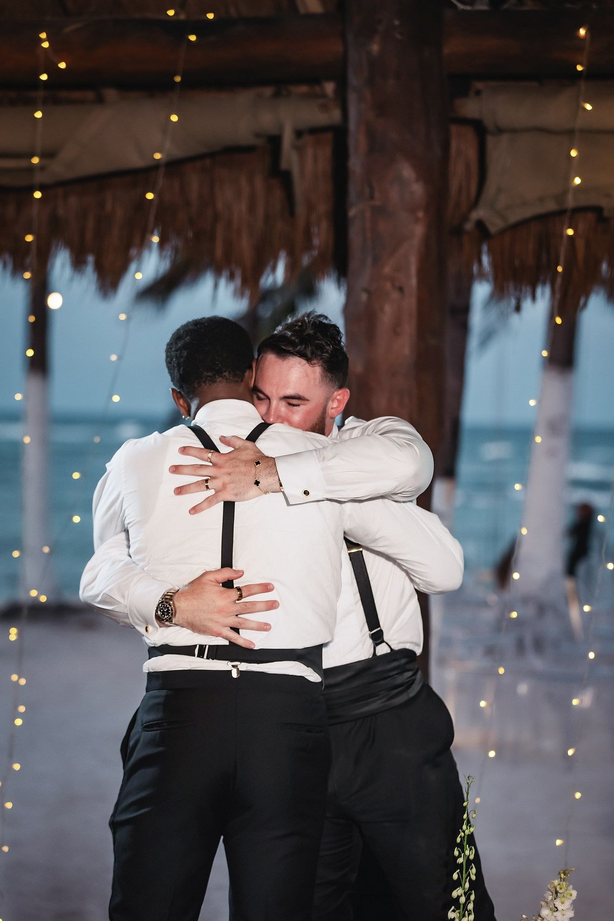 Two grooms sharing an emotional hug at their beach wedding reception, surrounded by warm fairy lights.