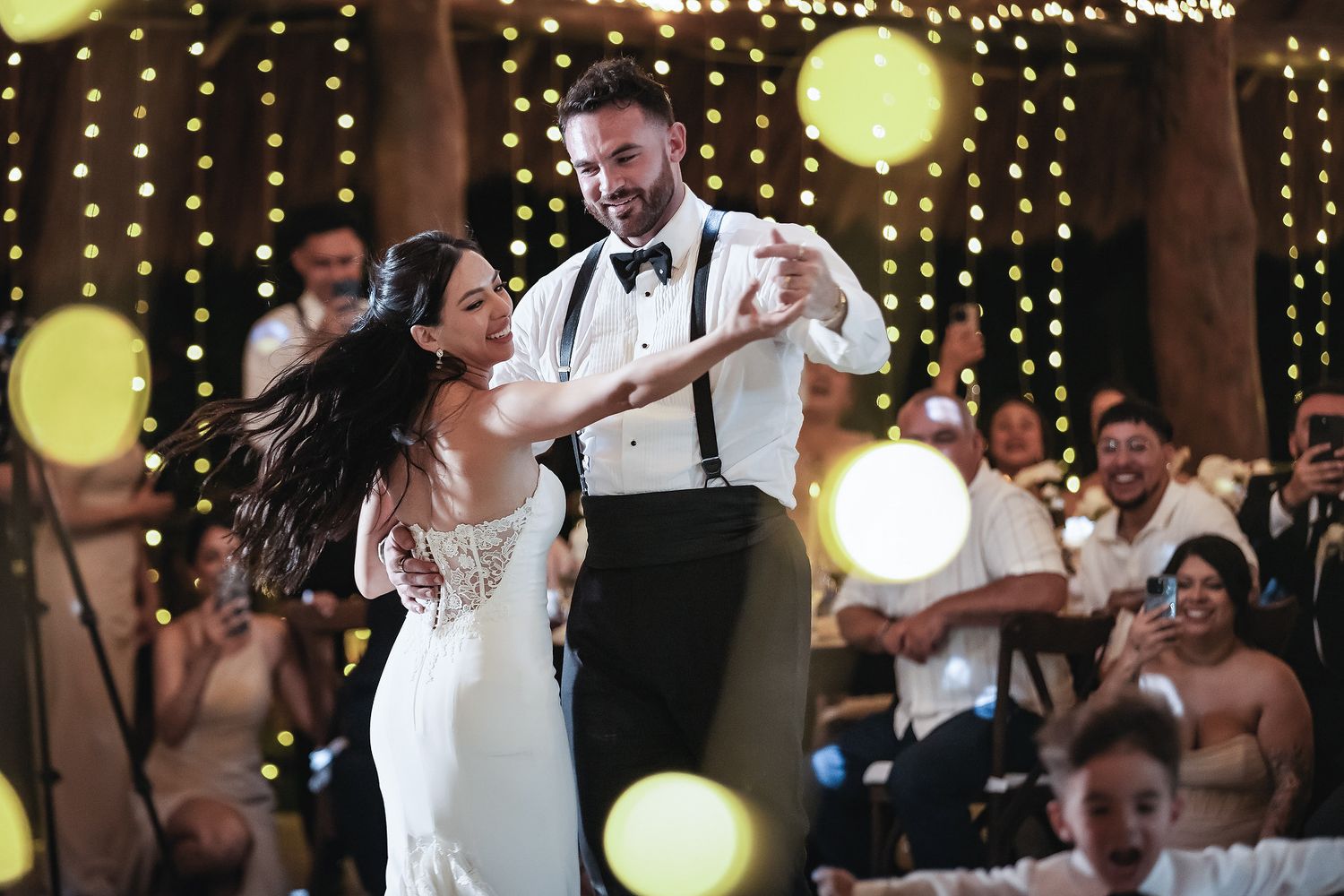 Bride and groom share their first dance at a romantic wedding reception with warm bokeh string lights glowing in the background.