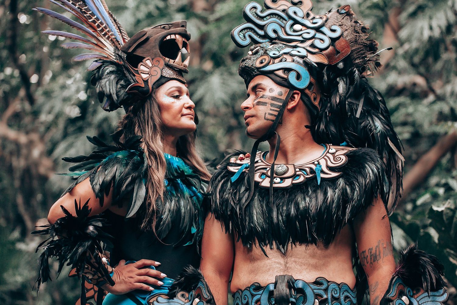 Two performers in stunning Mayan ceremonial costumes with feathers and face paint gaze at each other in the jungle.