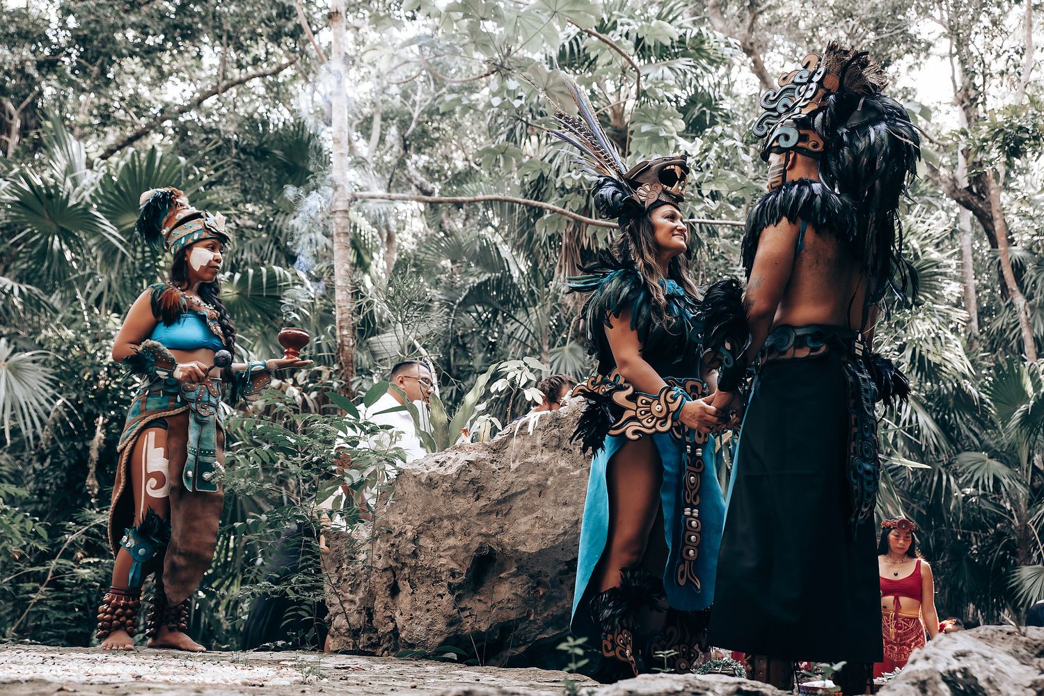 Three performers in traditional Mayan attire with turquoise and black feathered costumes stand in a jungle clearing.