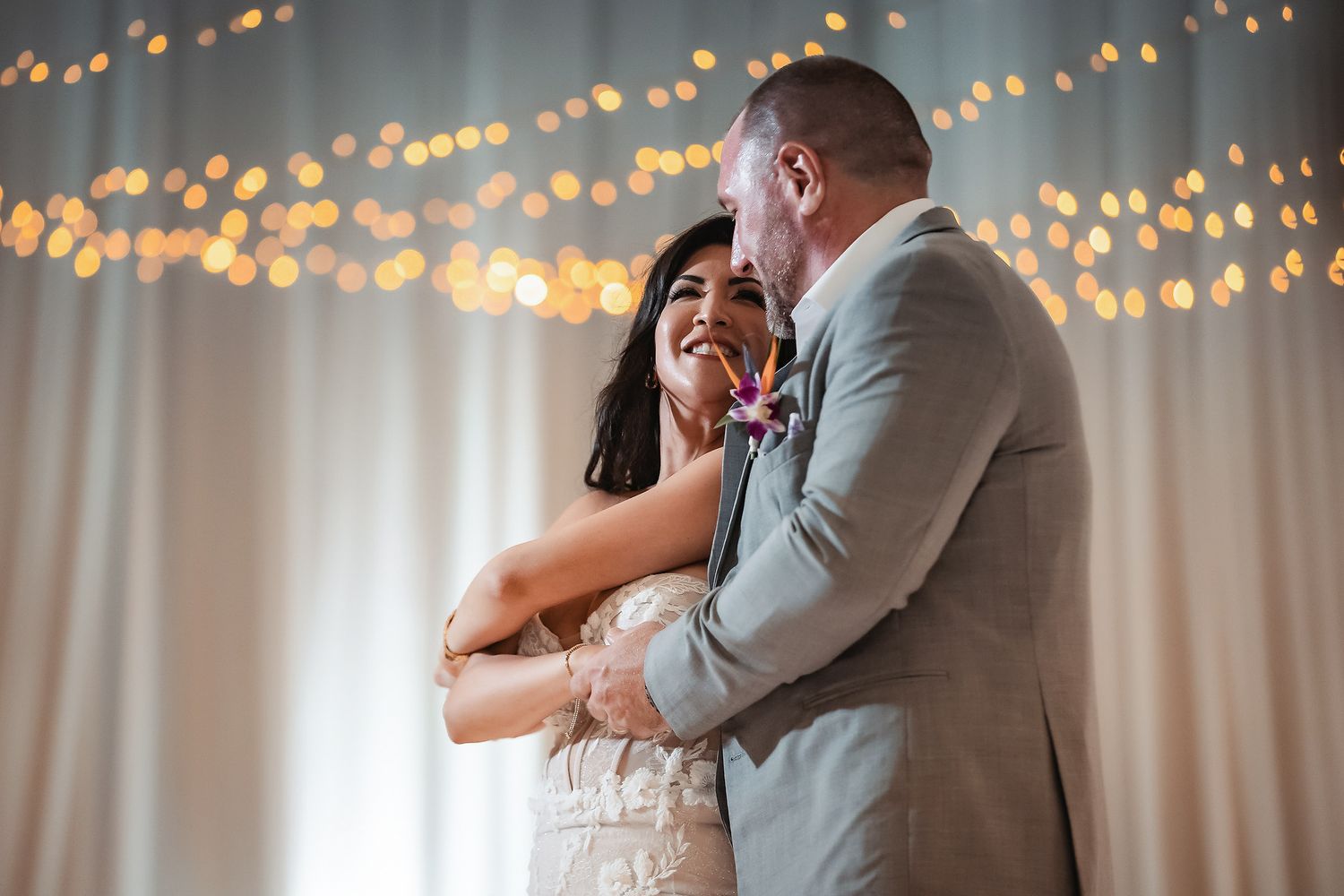 Bride and groom share first dance, smiling joyfully, with warm bokeh string lights glowing behind white drape backdrop.