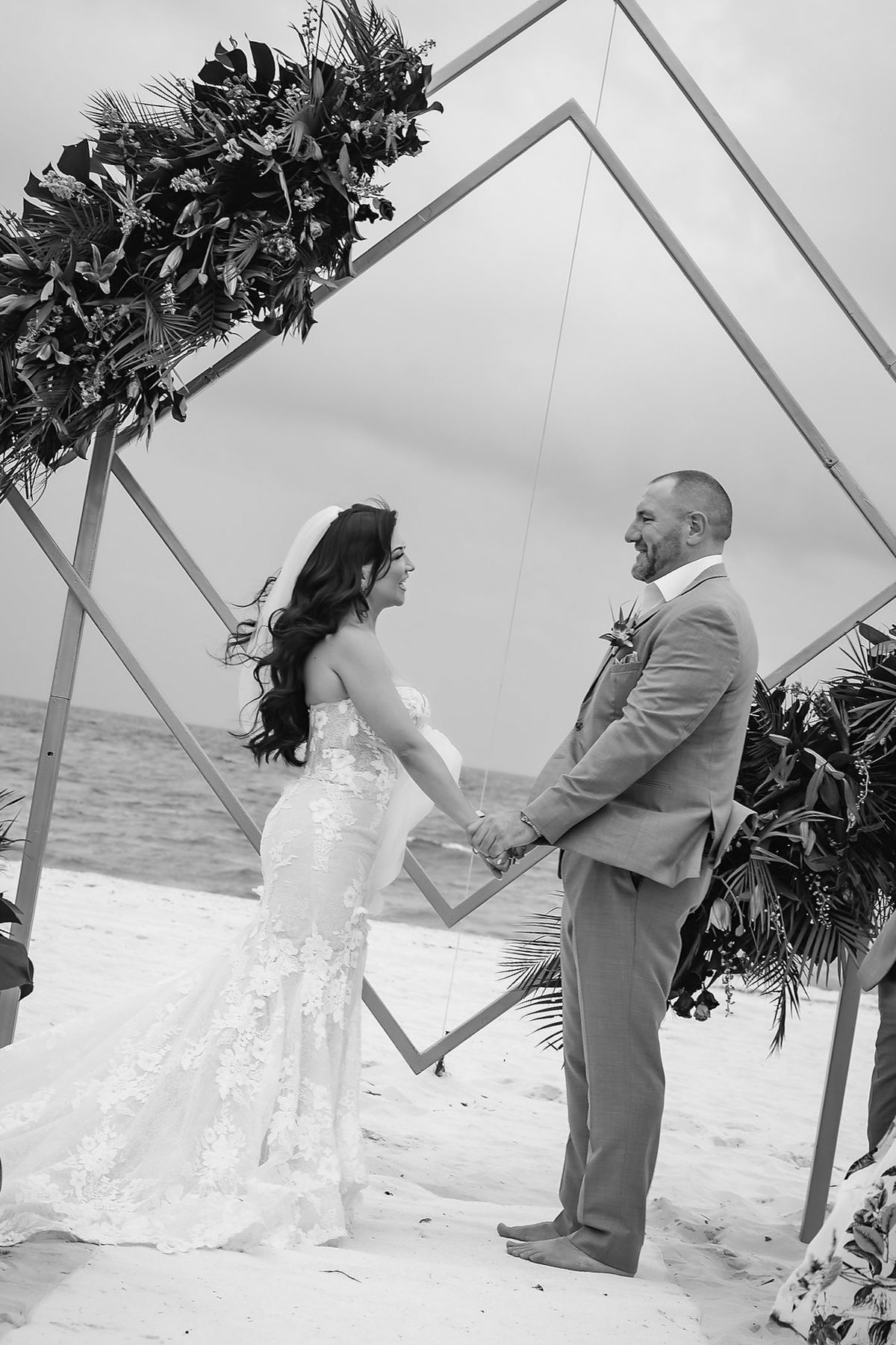 Bride and groom holding hands at beach wedding ceremony under geometric arch with tropical floral arrangements.