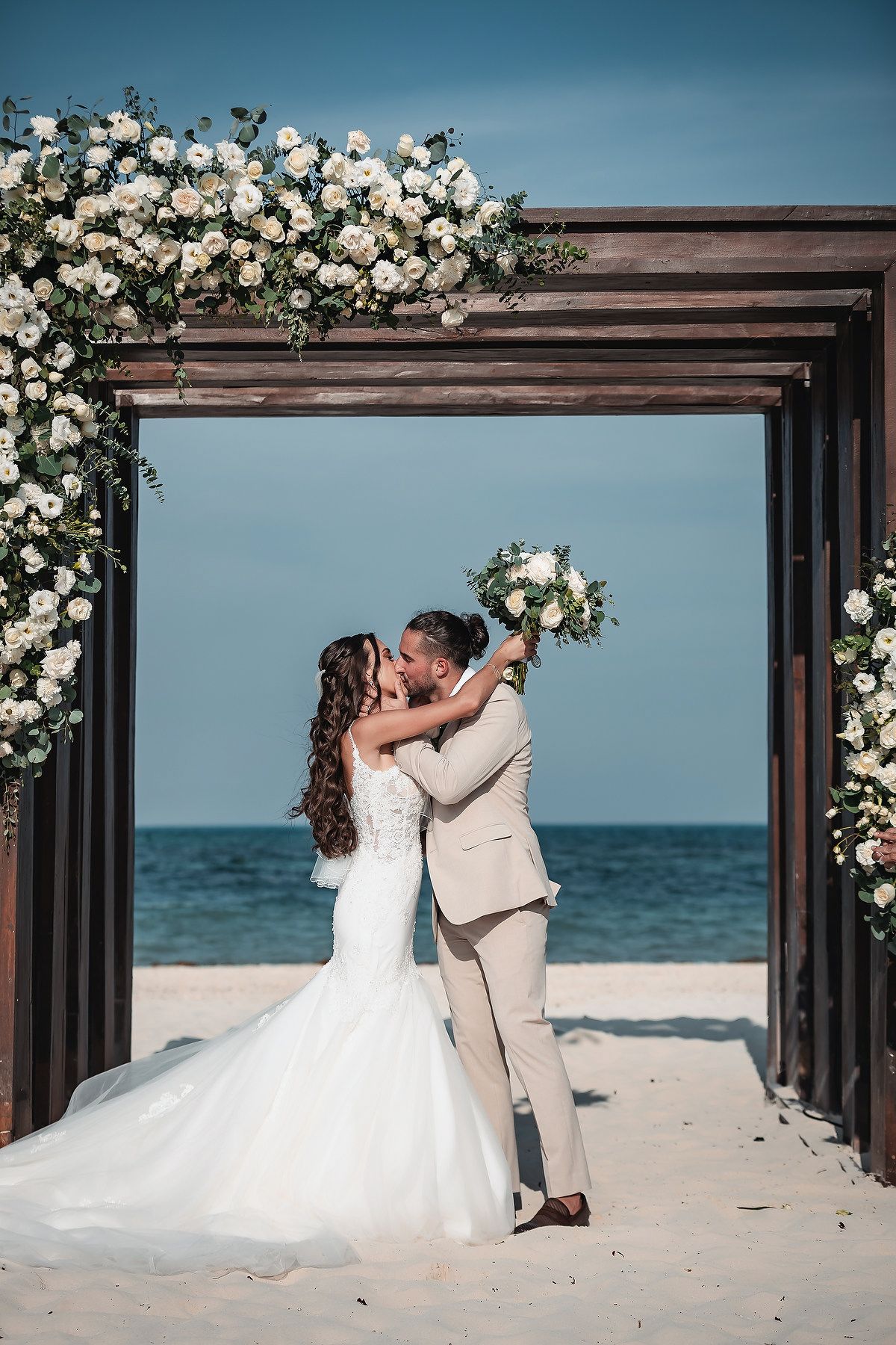 Bride and groom kissing under a floral arch on a beach, with white roses and ocean views in the background.