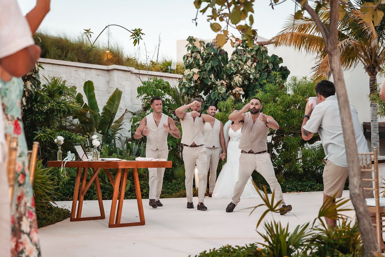 Groomsmen in tan vests celebrate outdoors at a tropical wedding venue with lush greenery and palm trees.