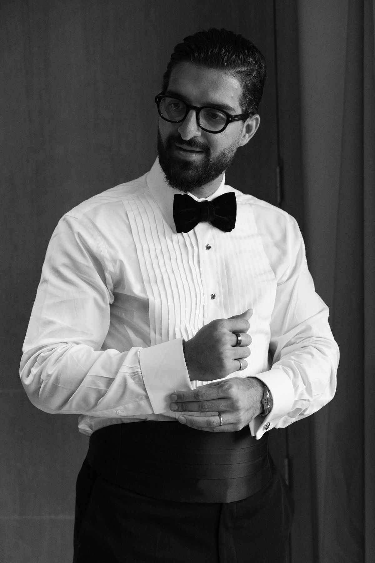Dapper bearded groom in white tuxedo shirt and bow tie fastening cufflinks, elegant black and white photo.