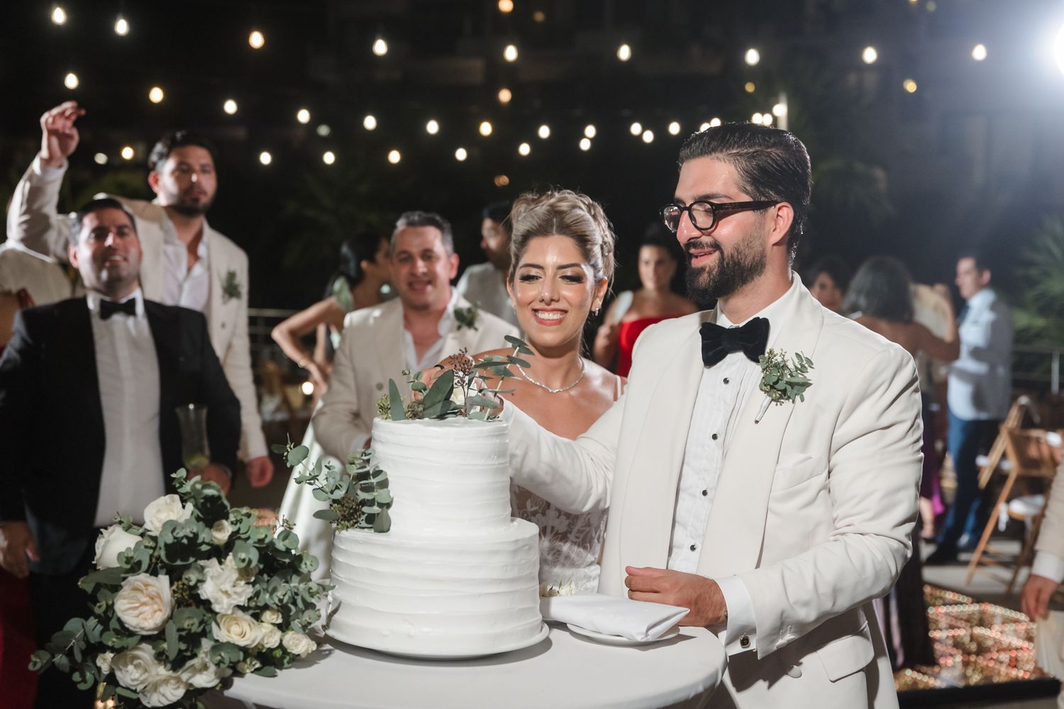 Bride and groom cutting white wedding cake at elegant outdoor reception with string lights.