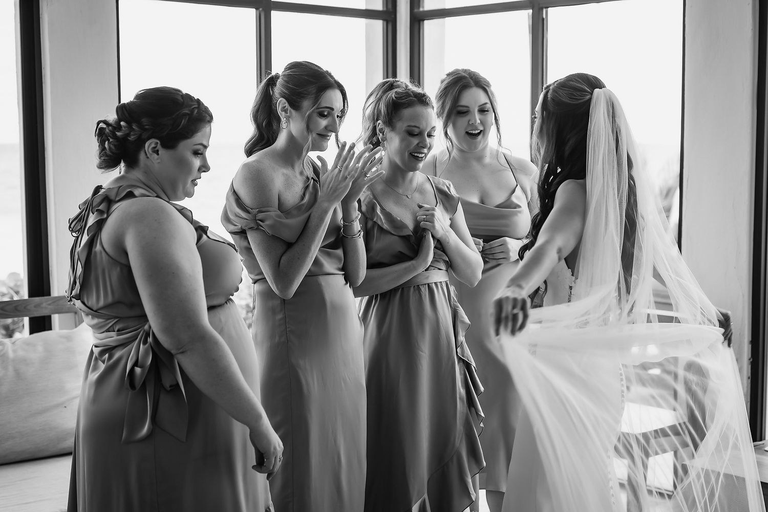 Bridesmaids in matching dresses react with joy as bride in veil shows her wedding look in black and white photo.