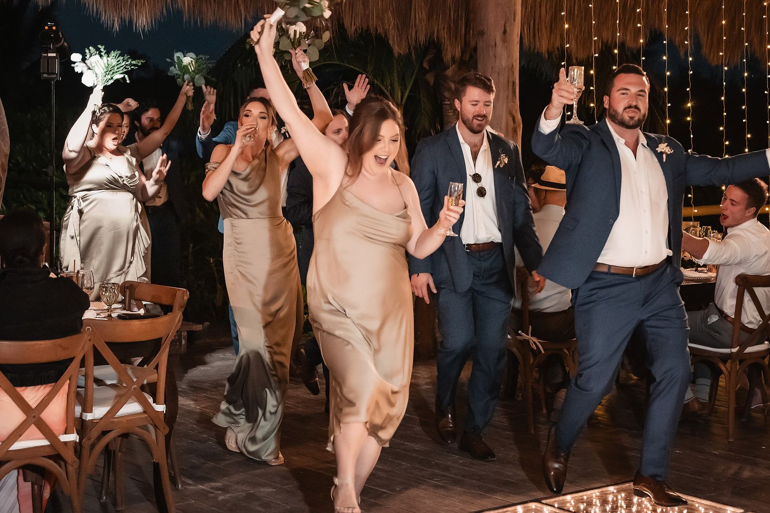 Wedding guests in formal attire dancing joyfully at an outdoor reception with string lights.