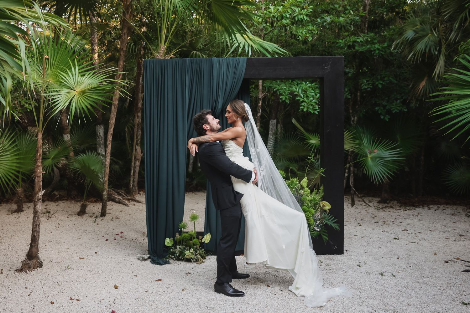 Bride and groom share romantic embrace outdoors under dark floral arch with teal draping surrounded by tropical greenery.
