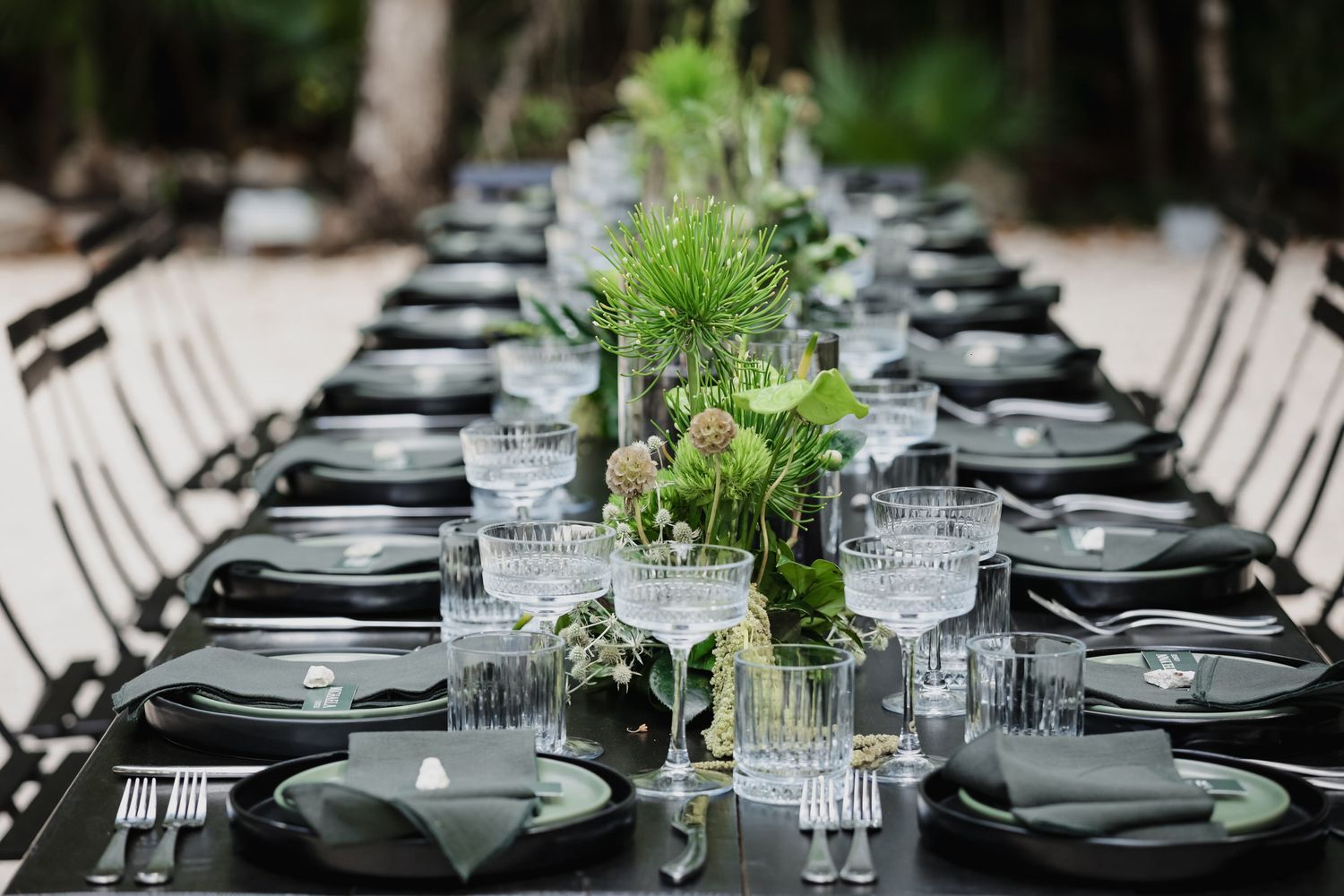 Elegant black long dining table with green botanical centerpieces, crystal glassware, and dark napkins for an outdoor wedding.