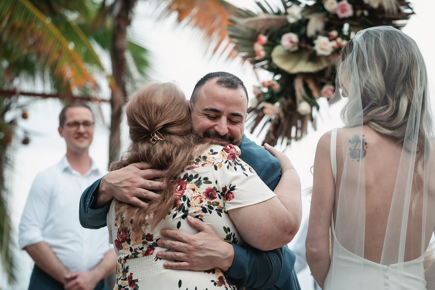 Groom in blue suit shares warm hug with wedding guest while bride in lace gown stands nearby at beach ceremony.