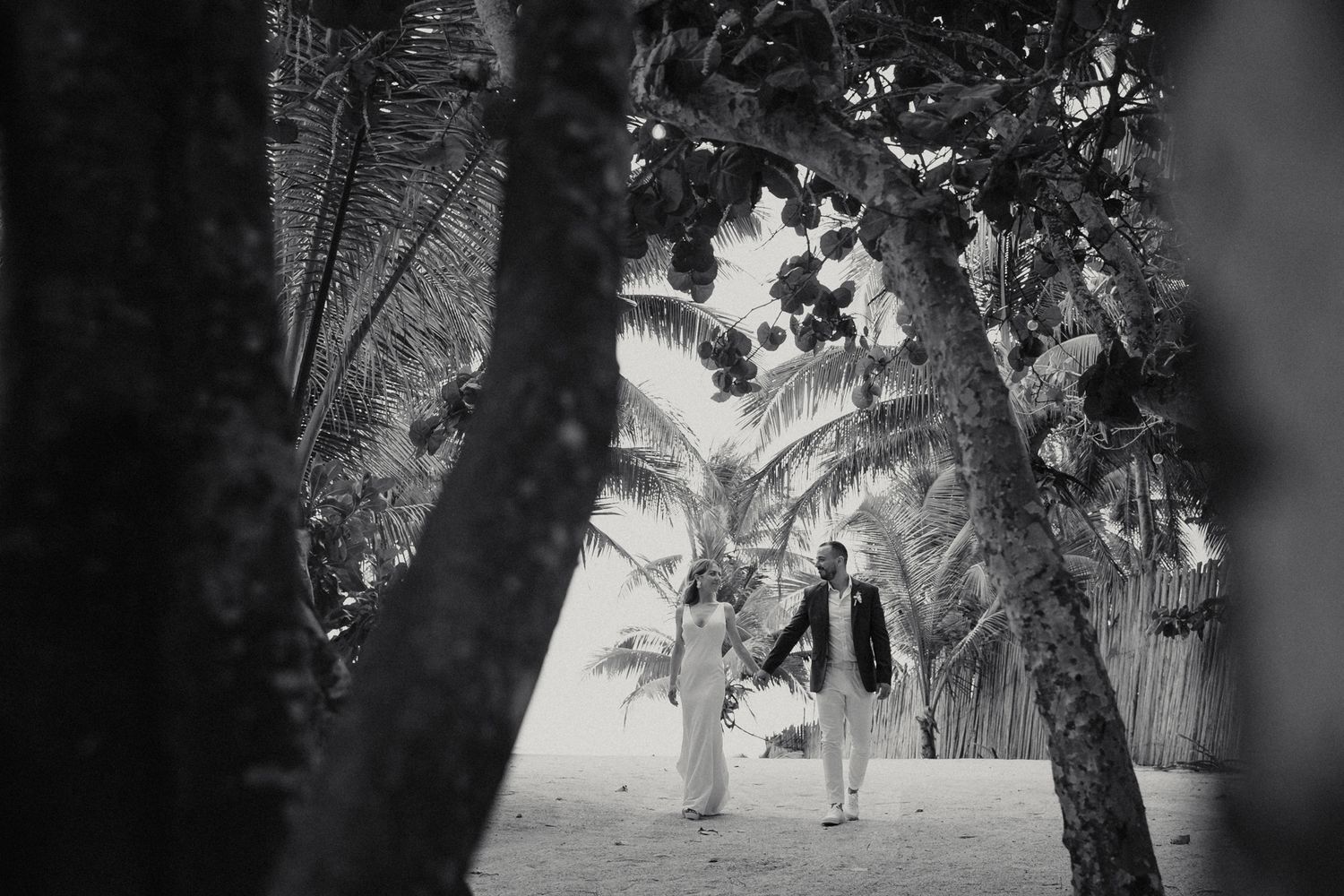 Black and white photo of bride and groom walking hand in hand between tropical palm trees.