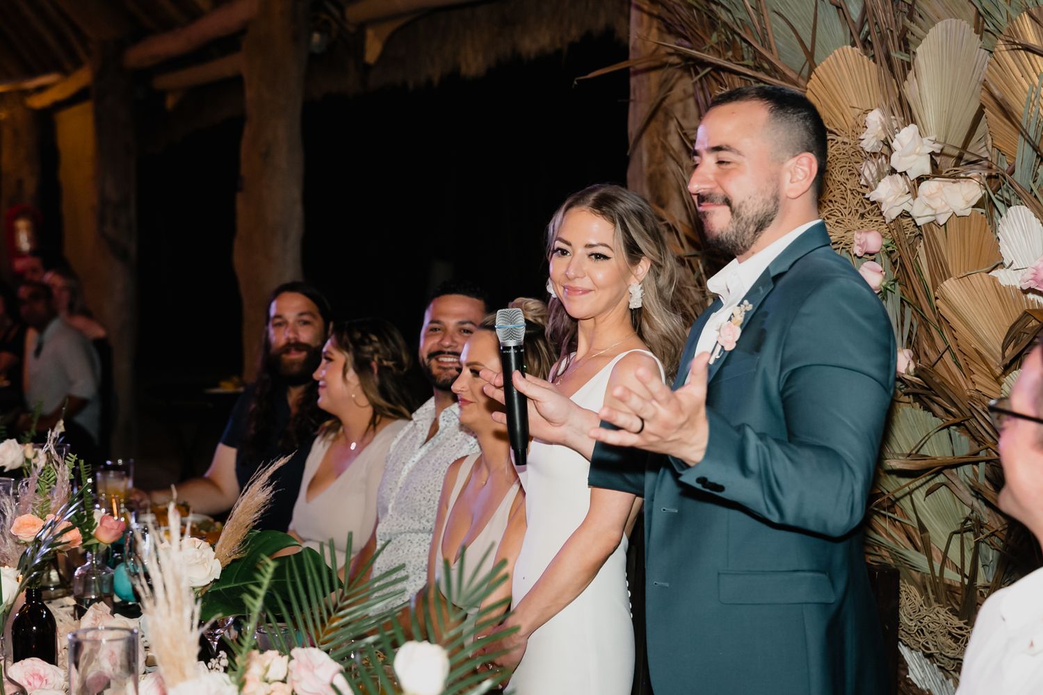 Bride and groom smile while speaking into a microphone at a tropical wedding reception with floral decor.