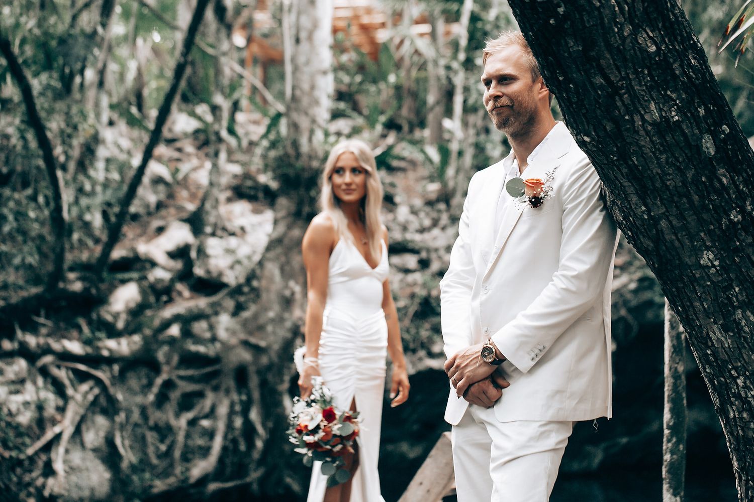Couple in white attire pose together in a tropical forest setting.