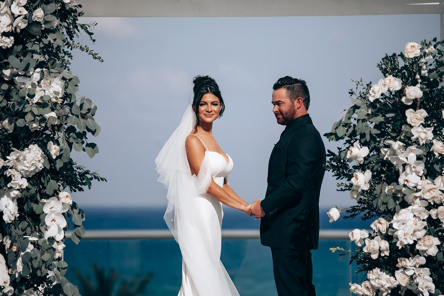 Romantic wedding portraits against ocean backdrop with white floral arch.