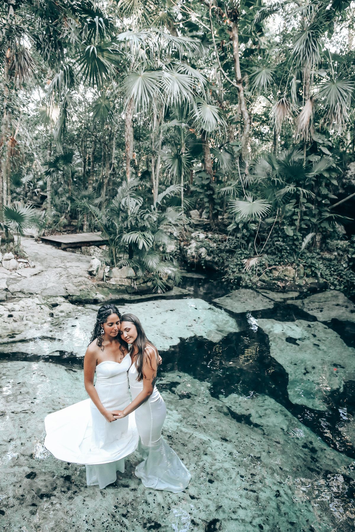 Couple in white dresses poses on rocky terrain surrounded by tropical forest vegetation.