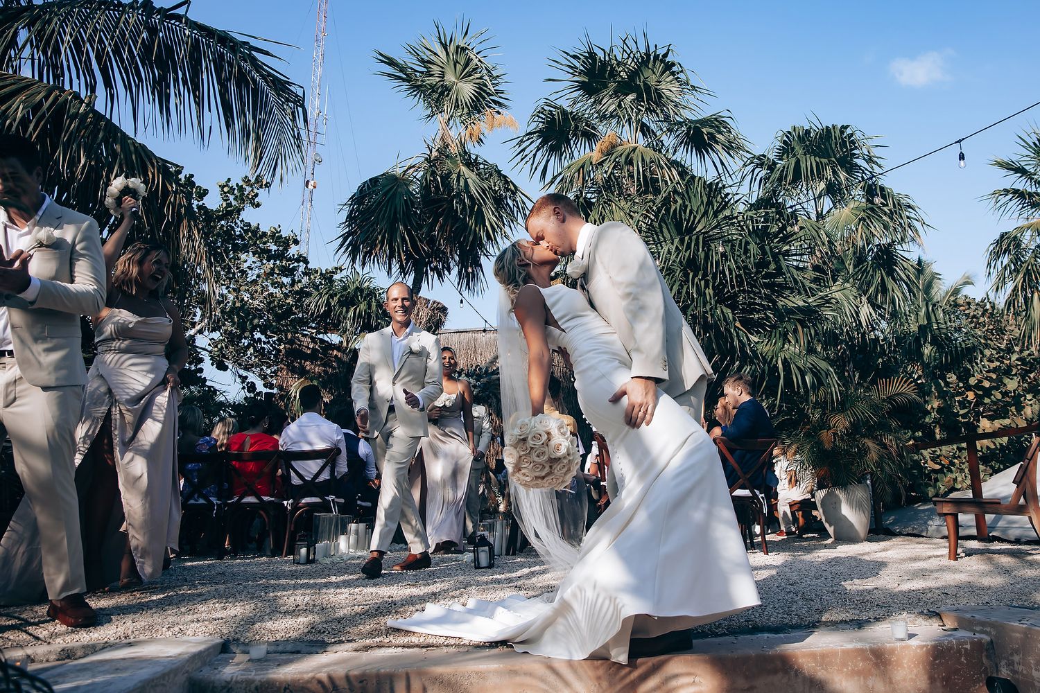 Outdoor wedding ceremony setup with palm trees and white chairs under bright sunlight.