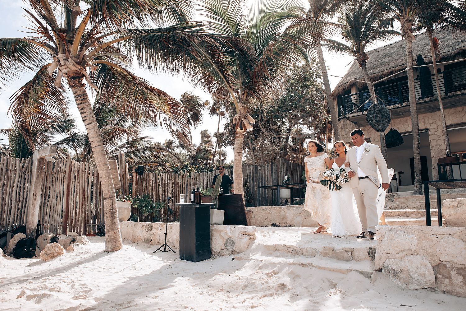 Beach wedding scene with palm trees and guests gathered along a sandy shoreline.