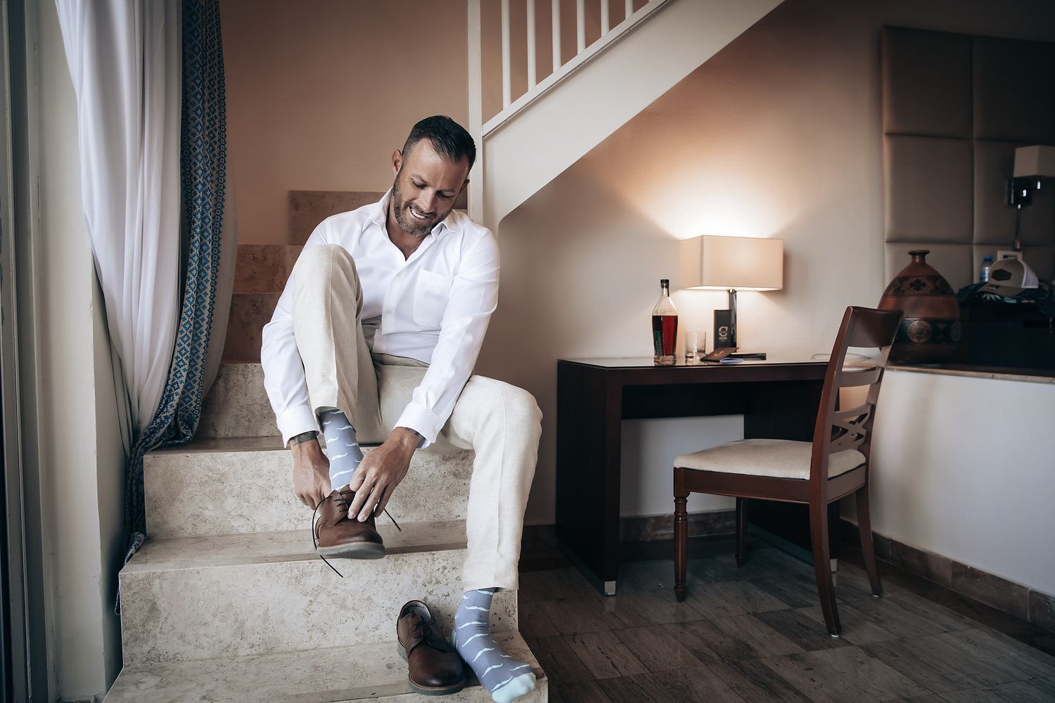 A man in a white suit sitting on a couch and putting on his shoes in a room.