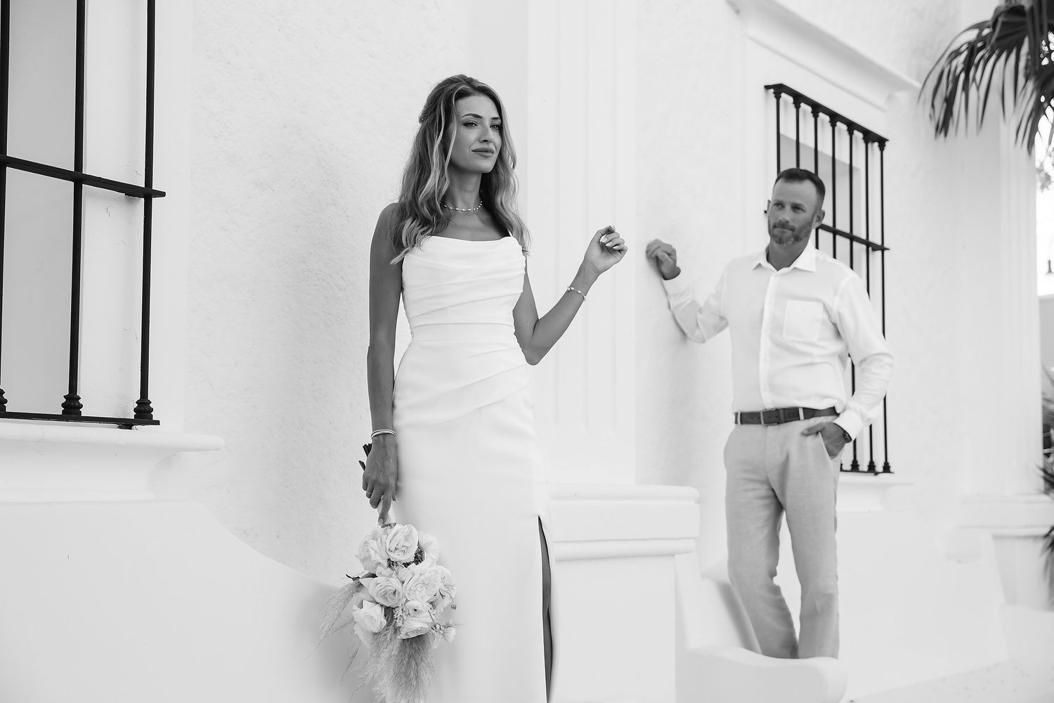 A bride in a white dress stands next to a groom in a suit, holding a bouquet of flowers.
