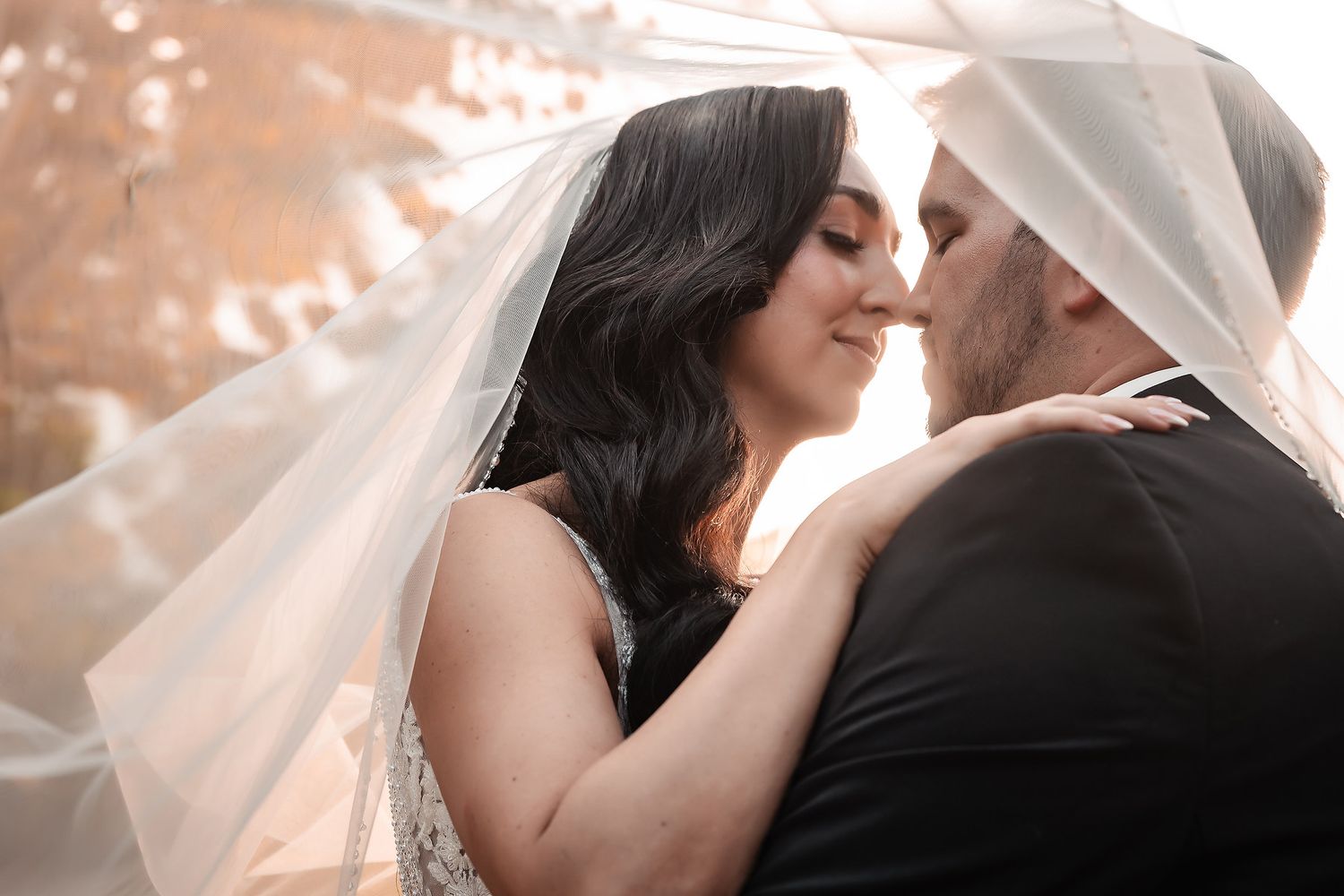 Bride and groom share a romantic moment under a flowing white veil at their wedding ceremony.
