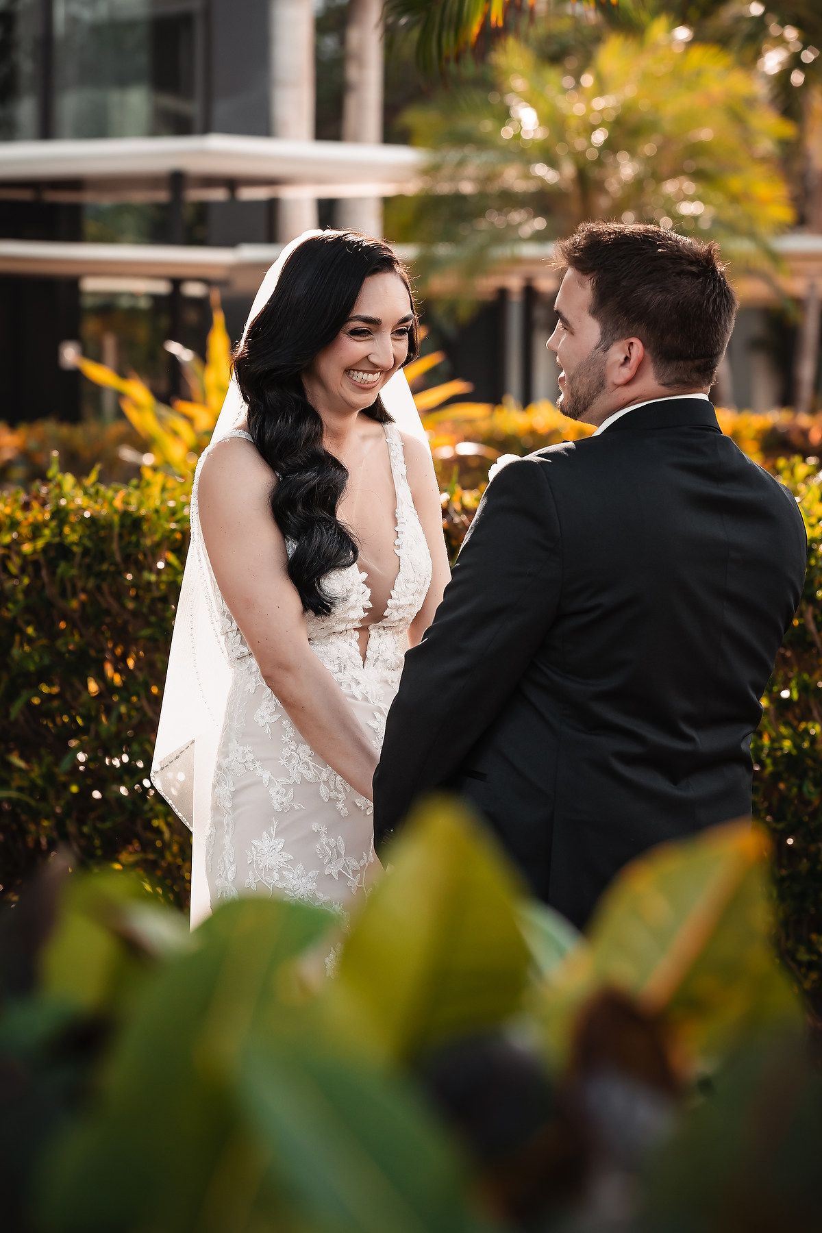 A smiling bride in a lace gown and veil shares a romantic moment with her groom in a suit outdoors.