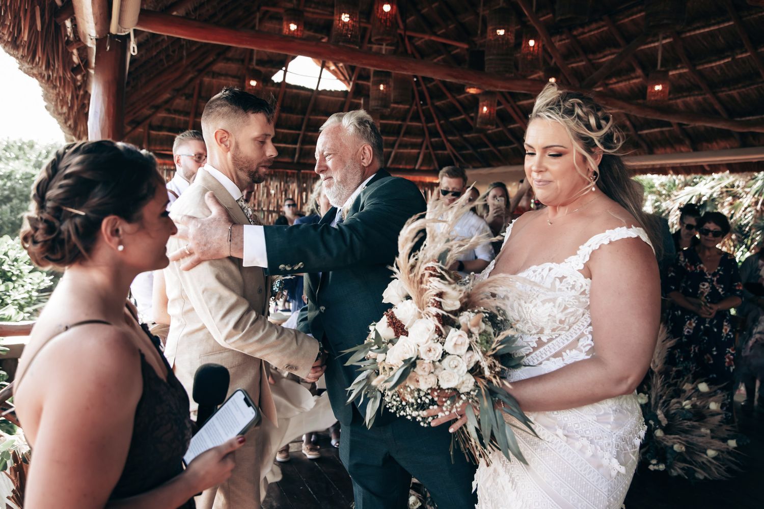 Bride in lace gown holding bouquet during outdoor wedding ceremony with groom and officiant under rustic pavilion.
