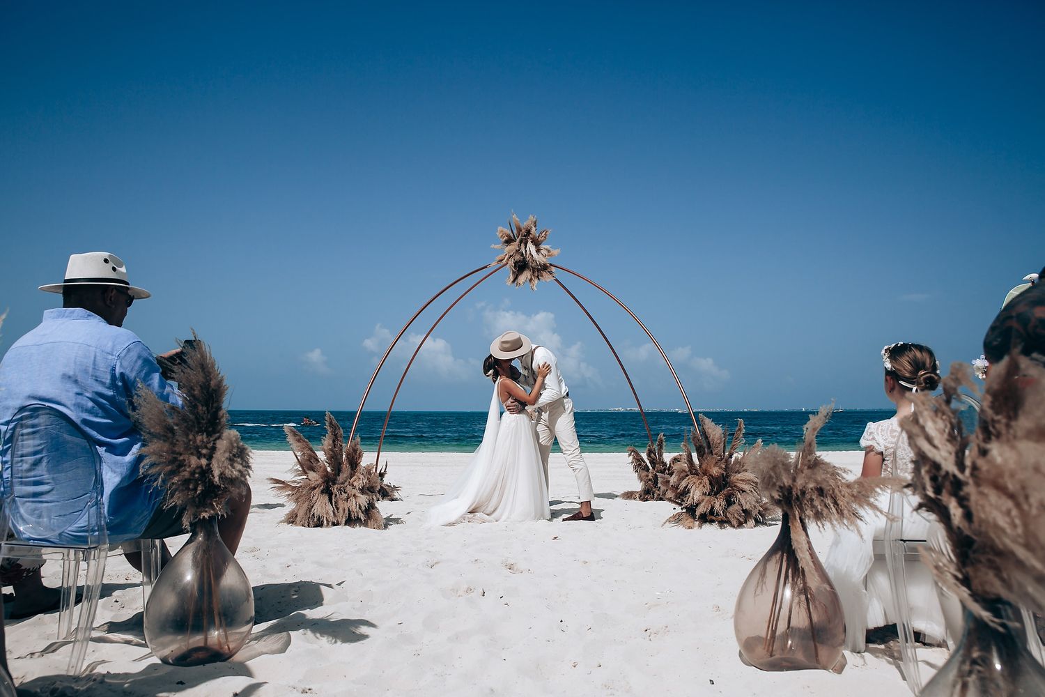 A beach wedding ceremony with dried palm decorations and guests watching under blue skies.