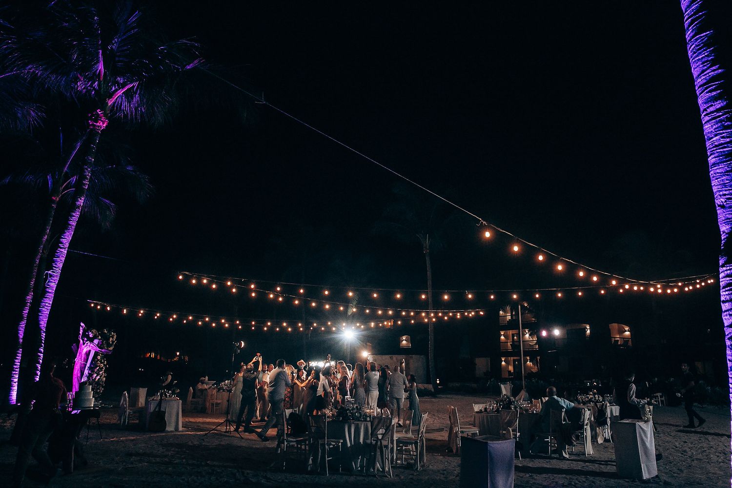 Evening beach wedding reception setup with string lights and purple-lit palm trees creating ambient atmosphere.