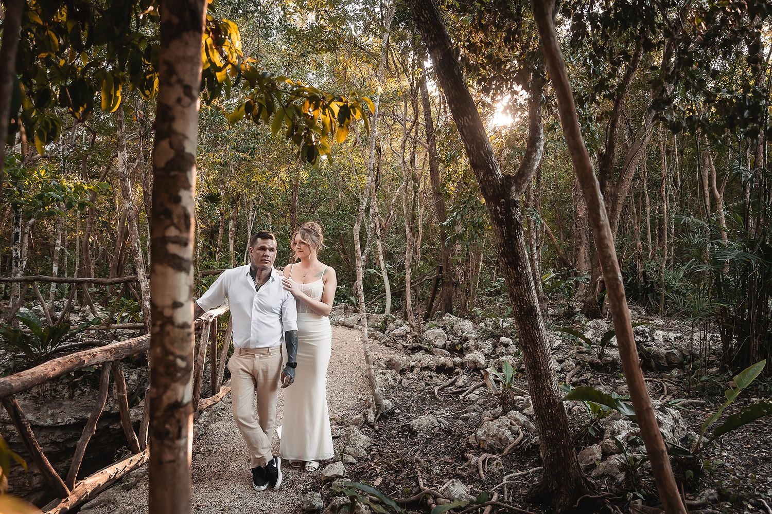 Couple in wedding attire walking hand-in-hand on a forest path surrounded by lush tropical trees.
