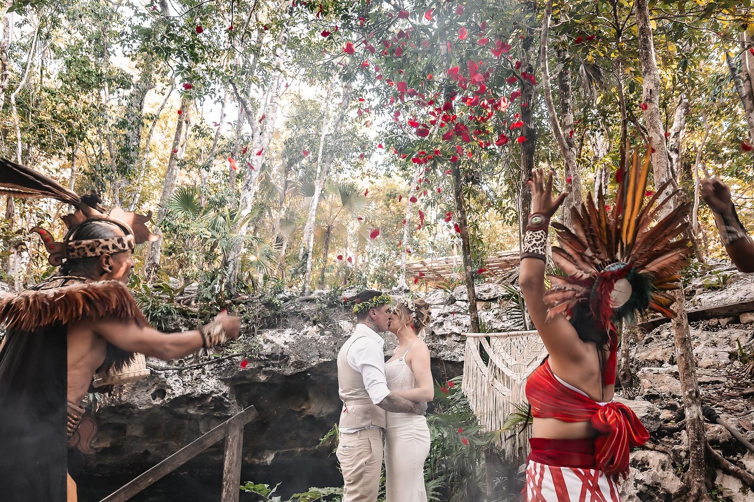 Couple exchanges vows in a lush jungle setting with Mayan performers in traditional headdresses and ceremonial attire.