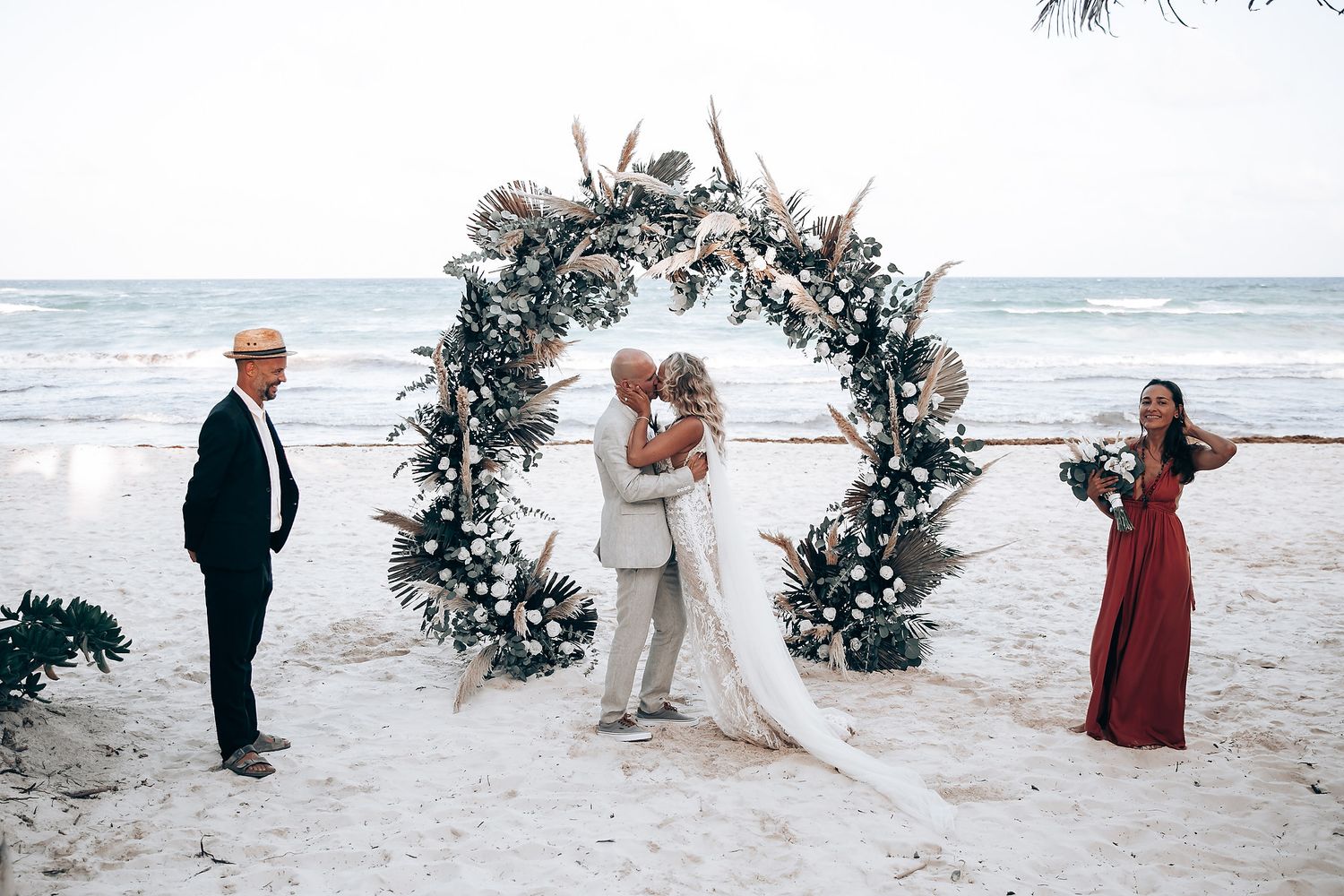 A beachside wedding ceremony features a large floral arch with guests watching the proceedings.