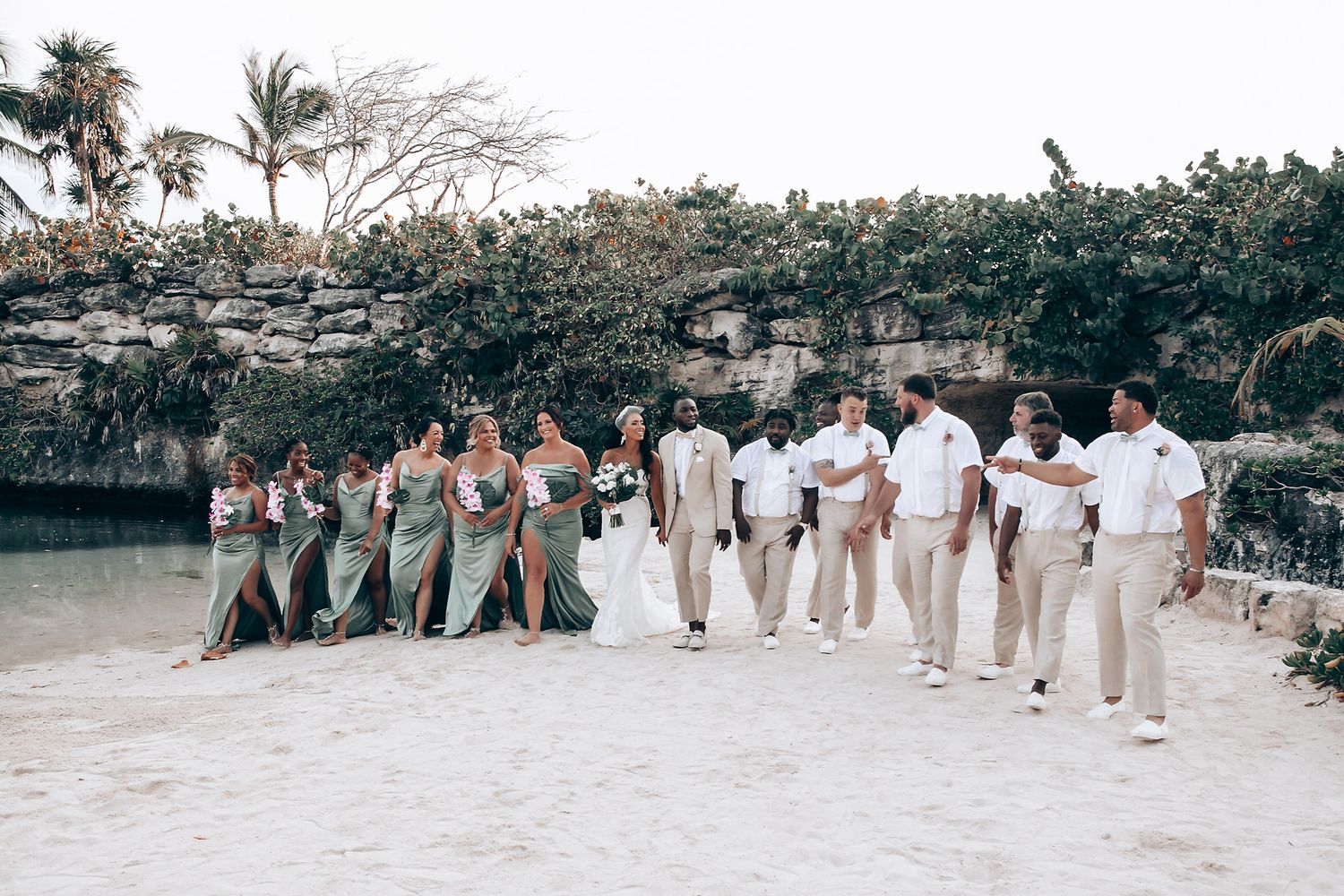 Wedding party poses on beach with bridesmaids in green and groomsmen in beige.