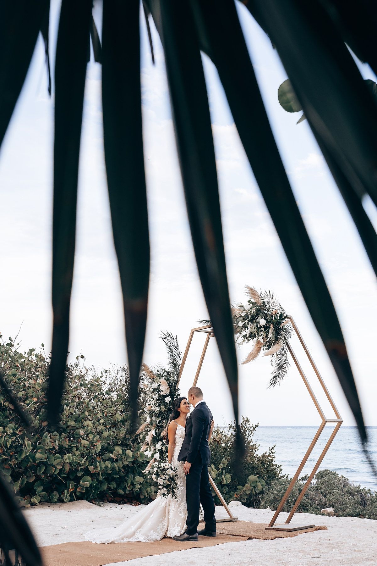 Beach wedding ceremony viewed through palm fronds with ocean backdrop.