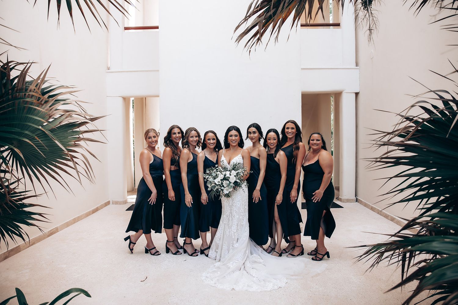 Wedding party in black dresses posing together against white wall with palm fronds.