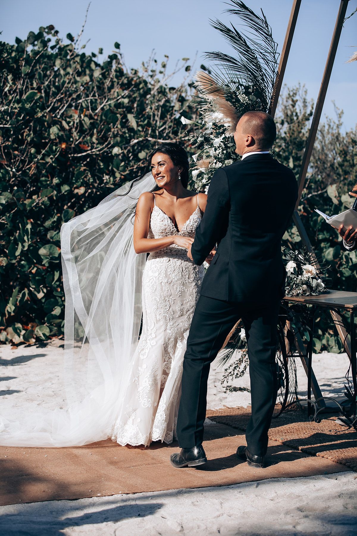 A couple exchanges vows during a tropical outdoor wedding ceremony with palm trees and greenery in the background.